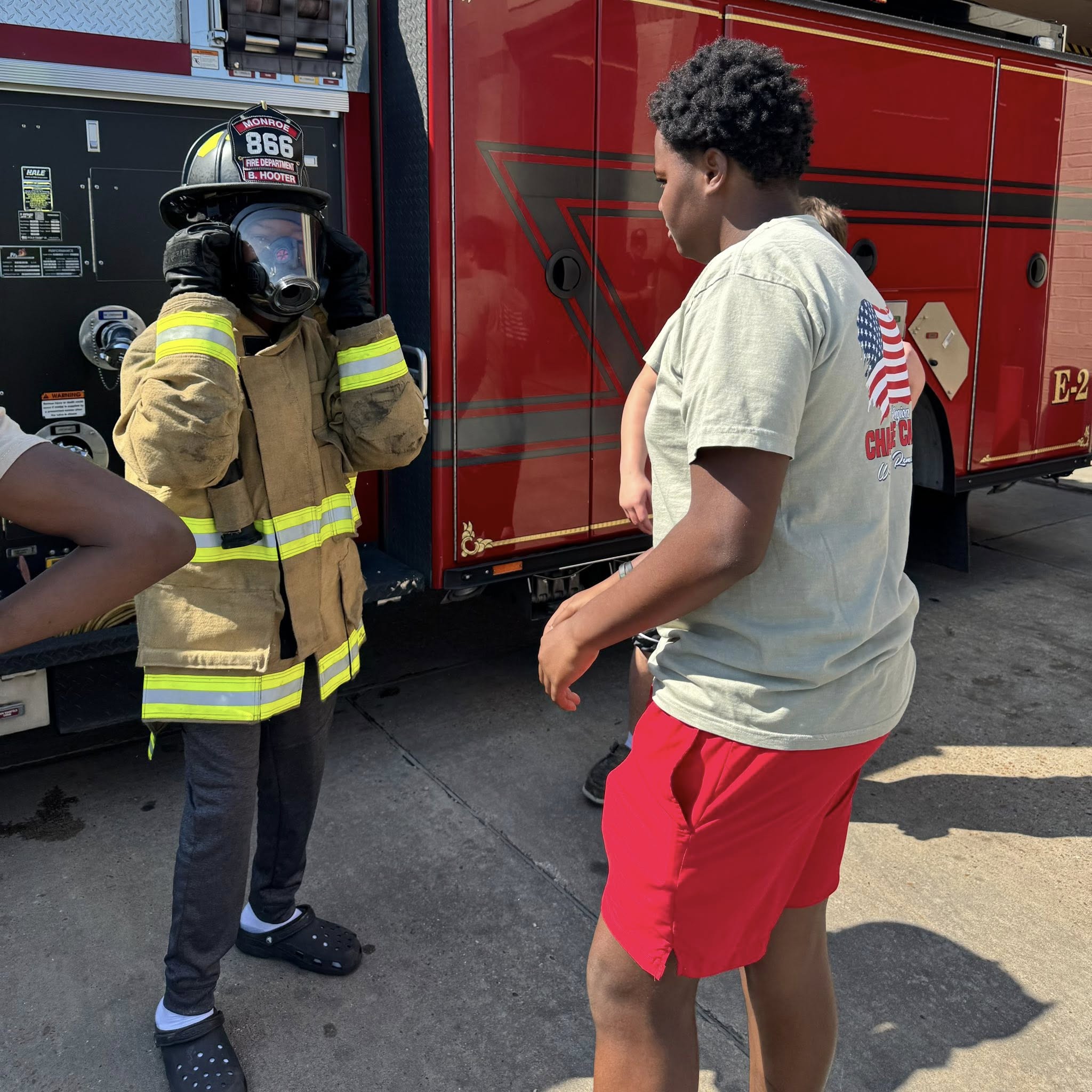 A boy looking at another kid wear full fireman's gear.